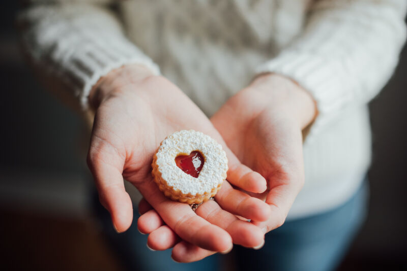 A woman in a white knitted sweater holding a Linzer cookie (with jam inside a heart cutout) in her crossed palms.